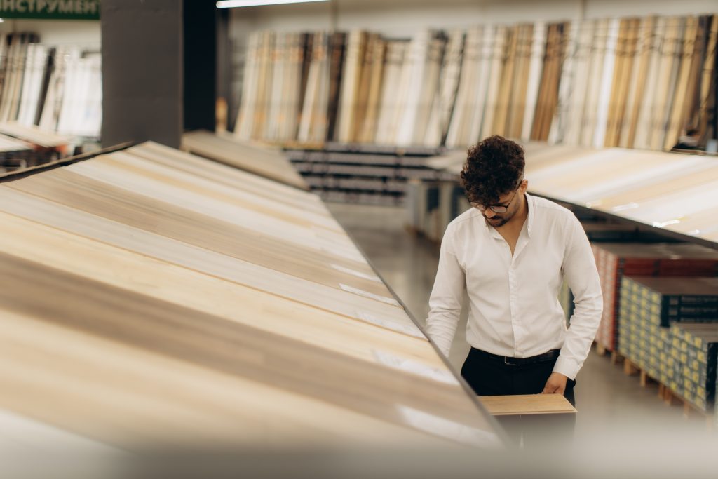 Neuer Fußboden - Parkett oder Laminat? 1 Ein Mann in einem weißen Hemd bewertet Holzpaneele in einem Baumarkt und vergleicht akribisch ihre Texturen und Oberflächen für ein Bodenbelagsprojekt. Reihenweise angezeigte Optionen sorgen für ein umfassendes Einkaufserlebnis.