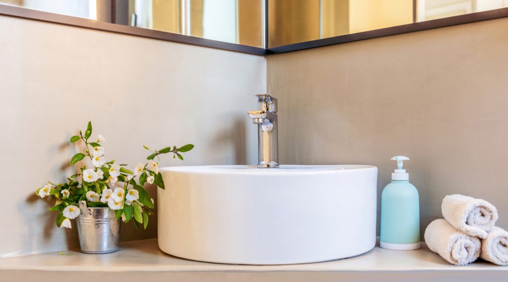 10 Tipps fürs Badezimmer 2 Waschbecken und Wasserhahn im Badezimmer, moderne Inneneinrichtung. Mischbatterie aus Chrom und rundem Porzellan-Waschbecken. Seifenspender, Handtücher und Blumentopf.