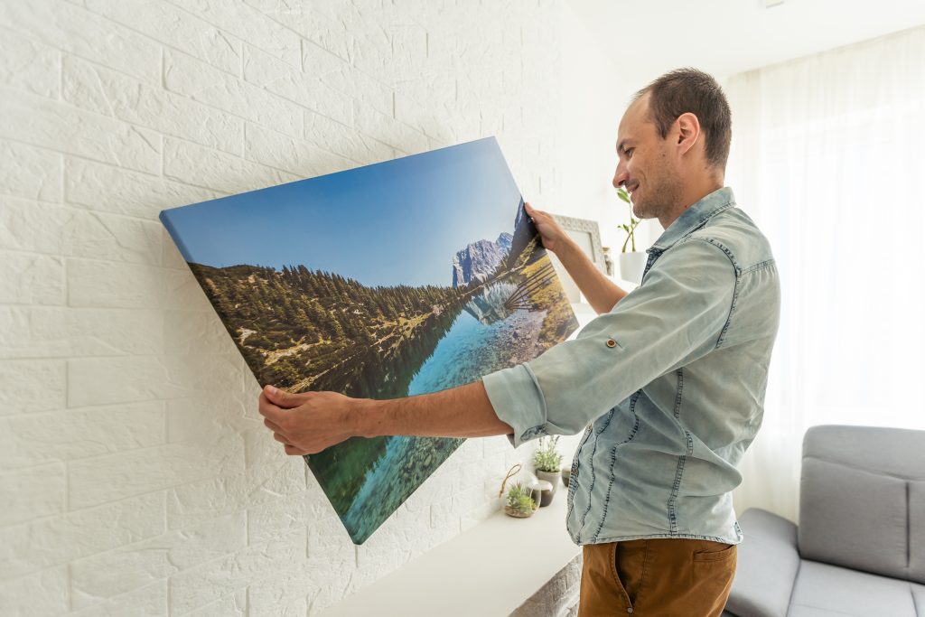 Die Verjüngungskur für das eigene Zuhause: 7 coole Ideen 2 Leinwanddruck mit Galeriefolie und Innendekor aus trockenem Gras. Frau hängt Fotografie an weiße Wand. Händehaltender Fotoleinwanddruck mit Bild einer Insel in der Stadt Biarritz in Frankreich. Foto in hoher Qualität