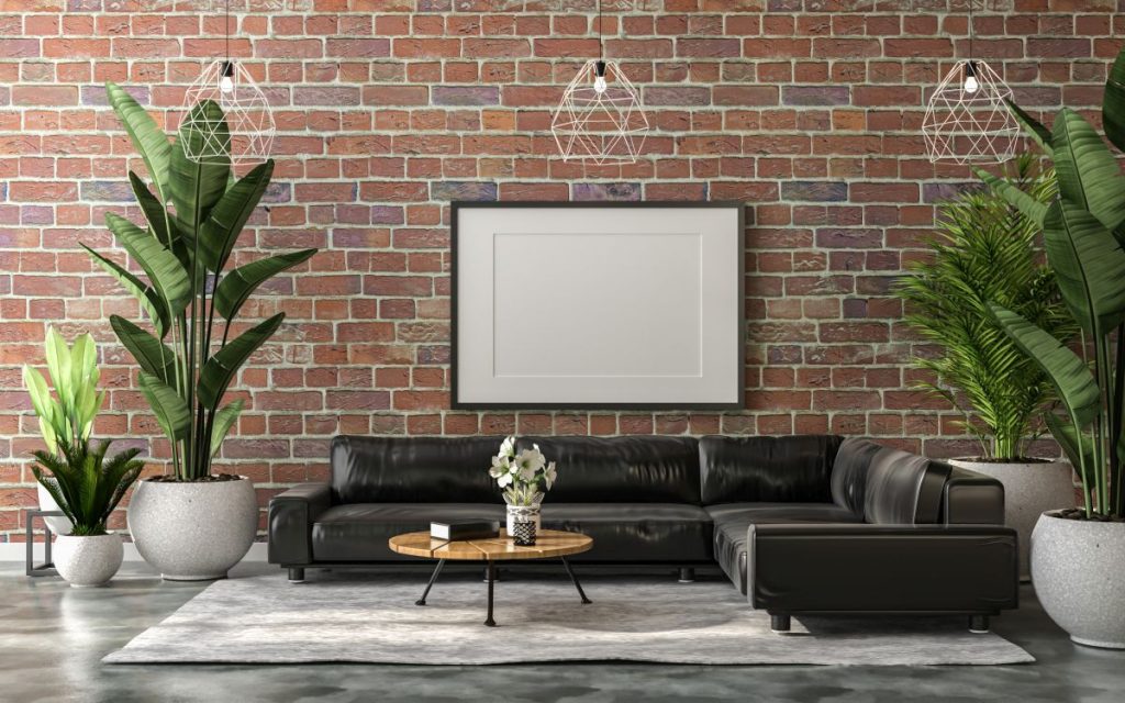 Industriell eingerichtetes Wohnzimmer mit roter Backsteinwand, grünen Zimmerpflanzen, einem dunkelgrauen Ecksofa und einem hellbraunen runden Holztisch auf einem weißen Teppich.
