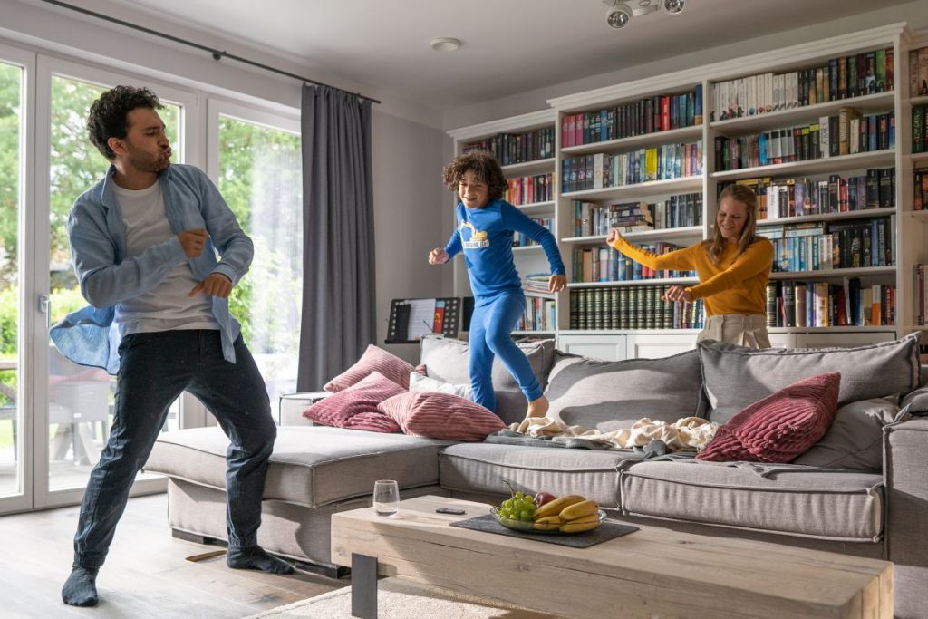 Eltern tanzen im Wohnzimmer, Kind hüpft auf Sofa herum