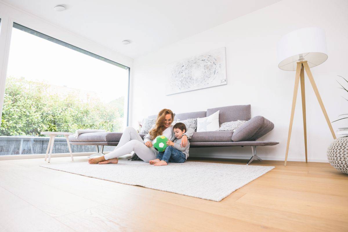 Mutter sitzt mit Sohn im WOhnzimmer auf einem Teppich. Im Hintergrund zu sehen: eine große Fensterfront und ein kleines Sofa