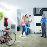 Kühlung Einfamilienhaus. Eine Familie steht mit Monteur lächelnd im Heizungskeller. Dort werden auch Fahrräder aufbewahrt. Der Vater hat das Kleinkind auf dem Arm.