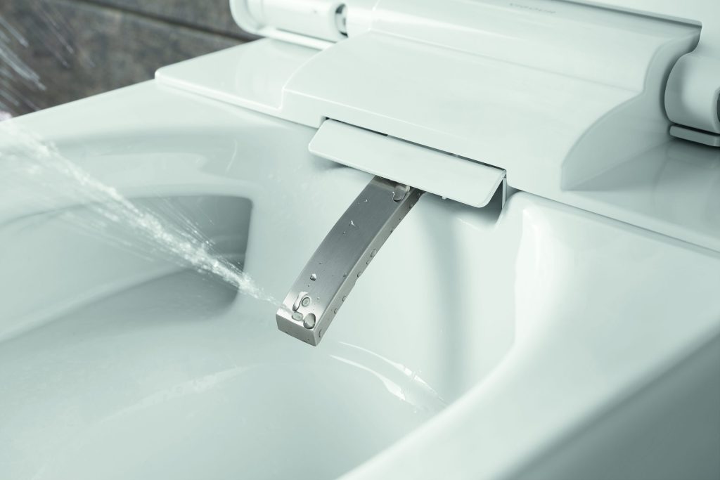 Nahaufnahme eines weißen Dusch-WCs mit ausgefahrenem Duscharm aus Edelstahl, aus dem ein präziser Wasserstrahl austritt.