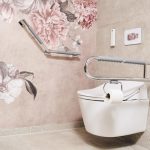 Barrierefreie Toilettenanlage: Weißes Wand-WC mit integriertem Toilettenpapierhalter am seitlichen, hochklappbaren Stützgriff. An der angrenzenden Wand befindet sich ein winkelbarer Haltegriff vor einer Tapete mit großflächigen, altrosa Pfingstrosen-Illustrationen. Die Fliesen sind in hellem Beige gehalten.