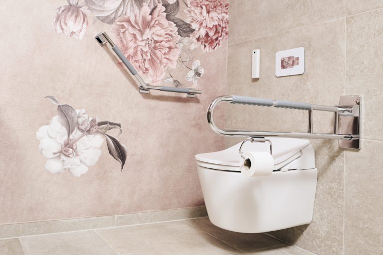 Barrierefreie Toilettenanlage: Weißes Wand-WC mit integriertem Toilettenpapierhalter am seitlichen, hochklappbaren Stützgriff. An der angrenzenden Wand befindet sich ein winkelbarer Haltegriff vor einer Tapete mit großflächigen, altrosa Pfingstrosen-Illustrationen. Die Fliesen sind in hellem Beige gehalten.