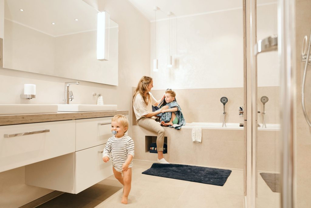 Ein großzügiges, hell beige gefliestes Badezimmer. Eine Frau sitzt am Rand einer eingebauten Badewanne und trocknet ein Kind mit einem Handtuch ab, während ein Kleinkind im gestreiften Body im Vordergrund läuft. Links befindet sich ein langer weißer Waschtisch mit großem Spiegel und modernen Chrom-Armaturen. Die Beleuchtung ist sanft und wohnlich.